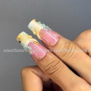 May include: Two long, square-shaped acrylic nails with a pink, white, and gold design. The nails feature a gradient effect, with gold and white at the tips and pink at the base. Embellishments include small gold and blue gems. The text "14nailsart" is visible.