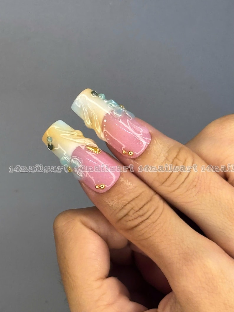 May include: Two long, square-shaped acrylic nails with a pink, white, and gold design. The nails feature a gradient effect, with gold and white at the tips and pink at the base. Embellishments include small gold and blue gems. The text "14nailsart" is visible.