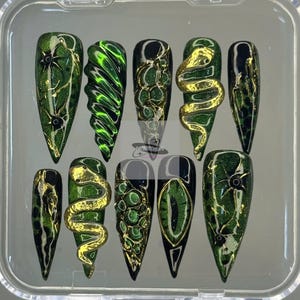 May include: A set of ten artificial nails in a clear plastic case. The nails feature a variety of designs, including green and gold glitter, gold snakes, and an eye design. The nails are long and pointed.