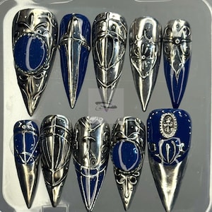 Puede incluir: Un conjunto de diez uñas postizas largas y puntiagudas. Cada uña presenta un diseño metálico plateado con detalles intrincados y un acento azul oscuro. Las uñas están dispuestas en una caja de plástico transparente.