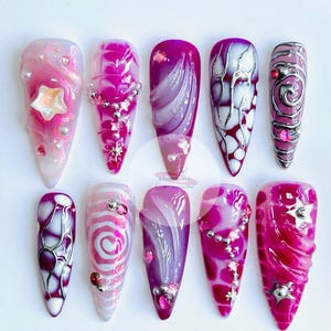 May include: A set of ten long, pointed artificial nails in shades of pink, purple, and white. The nails are decorated with glitter, beads, and star-shaped embellishments. Each nail has a unique design, with patterns and textures in a variety of styles.