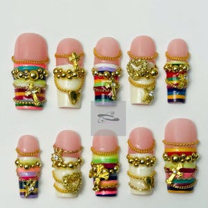 Op de afbeelding: Een set van tien kunstnagels met een roze basis en ingewikkelde gouden en juwelen versieringen. Elke nagel heeft een uniek ontwerp, waaronder kruisen, harten en kleurrijke strepen. De nagels zijn gerangschikt op een witte ondergrond.