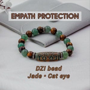 Könnte beinhalten: Ein Perlenarmband mit dem Text "EMPATH PROTECTION". Das Armband hat grünen Jade, braunes Tigerauge und eine zentrale DZI-Perle. Der Text "DZI bead, Jade, Cat eye" ist ebenfalls sichtbar.