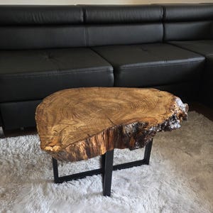 Könnte beinhalten: Rustikaler Couchtisch aus einem Baumstammquerschnitt gefertigt. Die natürliche Holzmaserung ist sichtbar, mit einer dunklen, strukturierten Kante. Der Tisch wird von einem schwarzen Metallrahmen getragen und schafft eine moderne, aber rustikale Ästhetik.