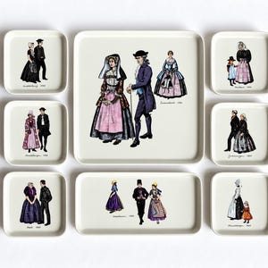 Puede incluir: Una colección de ocho bandejas de cerámica blanca de varios tamaños, cada una con ilustraciones coloridas de personas con vestimenta regional histórica. Los diseños incluyen etiquetas como Marken 1900, Hindeloopen 1900, Axel 1900, Zwanenburg 1900, Staphorst 1900, Scheveningen 1900 y Hindeloopen 1860.