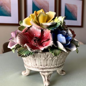 Könnte beinhalten: Ein keramisches Blumenarrangement in einer cremefarbenen Schale mit Füßen. Die Blumen sind in Gelb-, Rosa-, Blau- und Lilatönen gehalten, mit grünen Blättern. Die Schale hat ein strukturiertes, geripptes Design.