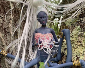 May include: A blue and white spider doll with long white hair, wearing a pink and white beaded top and gold bracelets. The doll is posed on a tree trunk.