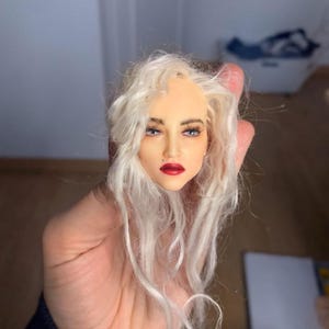 May include: A doll head with a pale complexion, red lips, and blue eyes. The doll head has long, wavy, white hair. The face is painted with detailed makeup. The doll head is held in a hand.