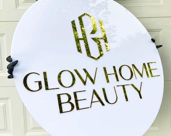 May include: A white round sign with gold lettering that reads "Glow Home Beauty" with a gold monogram of the letters "GH" in a geometric shape.