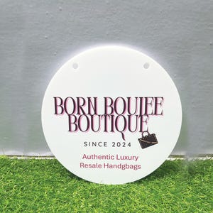 Op de afbeelding: Een wit rond bord met de tekst "BORN BOUJEE BOUTIQUE" in zwart met roze contouren. De tekst "SINCE 2024" staat onder de hoofdtekst, en de tekst "Authentic Luxury Resale Handbags" staat daaronder. Een klein beeld van een bruine handtas staat rechts van de tekst "SINCE 2024".