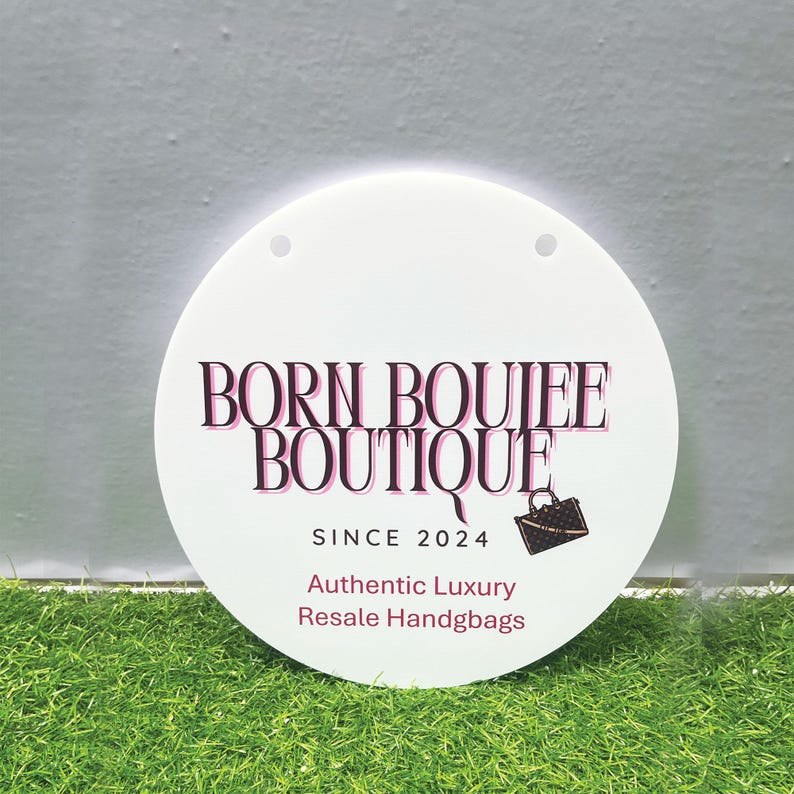 Op de afbeelding: Een wit rond bord met de tekst "BORN BOUJEE BOUTIQUE" in zwart met roze contouren. De tekst "SINCE 2024" staat onder de hoofdtekst, en de tekst "Authentic Luxury Resale Handbags" staat daaronder. Een klein beeld van een bruine handtas staat rechts van de tekst "SINCE 2024".