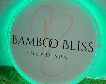 Puede incluir: Un letrero redondo blanco con un borde de neón verde. El letrero tiene un diseño de corazón estilizado y el texto "Bamboo Bliss - Head Spa -"