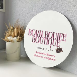 Op de afbeelding: Een wit rond bord met de tekst "Born Boujee Boutique Since 2024" in roze en zwarte letters. Het bord bevat ook de tekst "Authentic Luxury Resale Handbags" en een klein beeld van een bruine handtas.