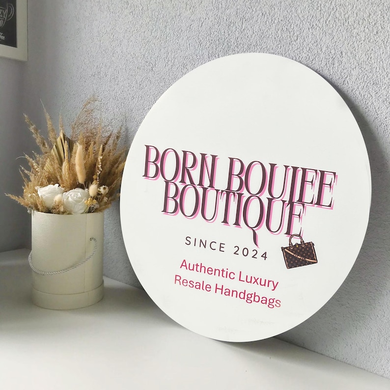 Op de afbeelding: Een wit rond bord met de tekst "Born Boujee Boutique Since 2024" in roze en zwarte letters. Het bord bevat ook de tekst "Authentic Luxury Resale Handbags" en een klein beeld van een bruine handtas.
