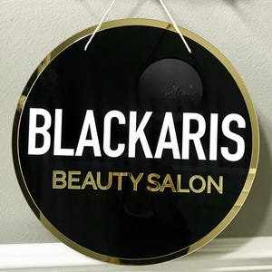Op de afbeelding: Rond zwart en gouden bord met de tekst "BLACKARIS BEAUTY SALON" in witte en gouden letters.