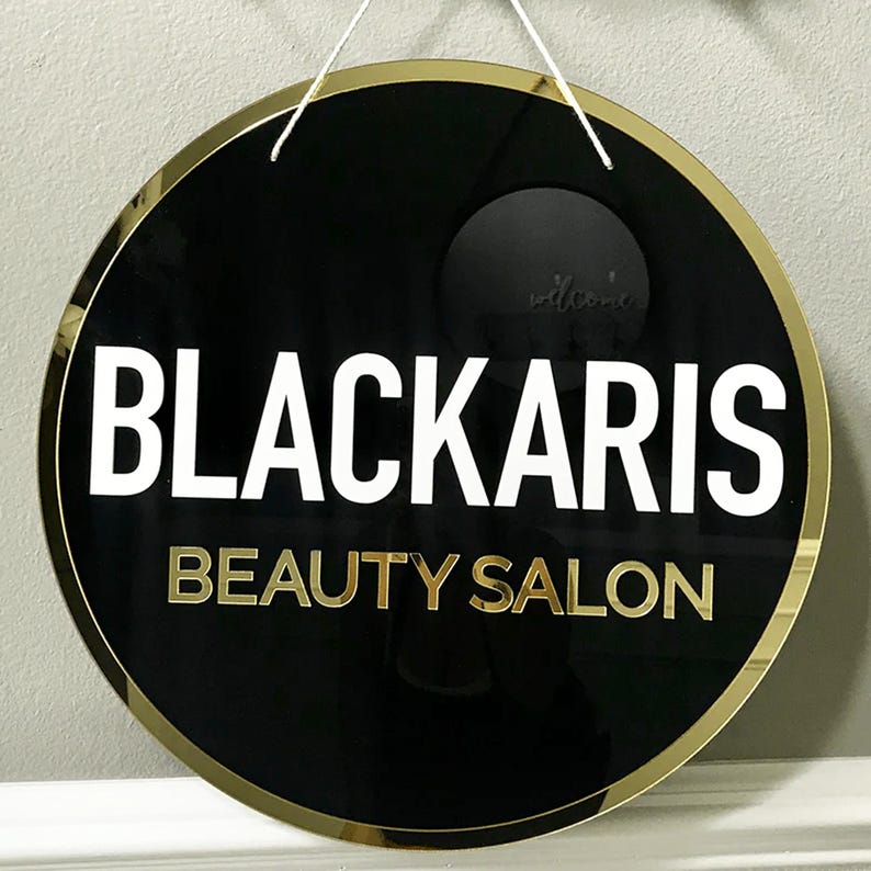 Op de afbeelding: Rond zwart en gouden bord met de tekst "BLACKARIS BEAUTY SALON" in witte en gouden letters.