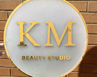 May include: A round, white sign with a gold border and the letters "KM" in gold, followed by the text "BEAUTY STUDIO" in gold.