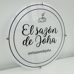 Op de afbeelding: Een wit rond bord met zwarte tekst en een afbeelding van een stuk kaas met een honinglepeltje. De tekst luidt "El saz&oacute;n de Joha" en "@elsazondejoha".