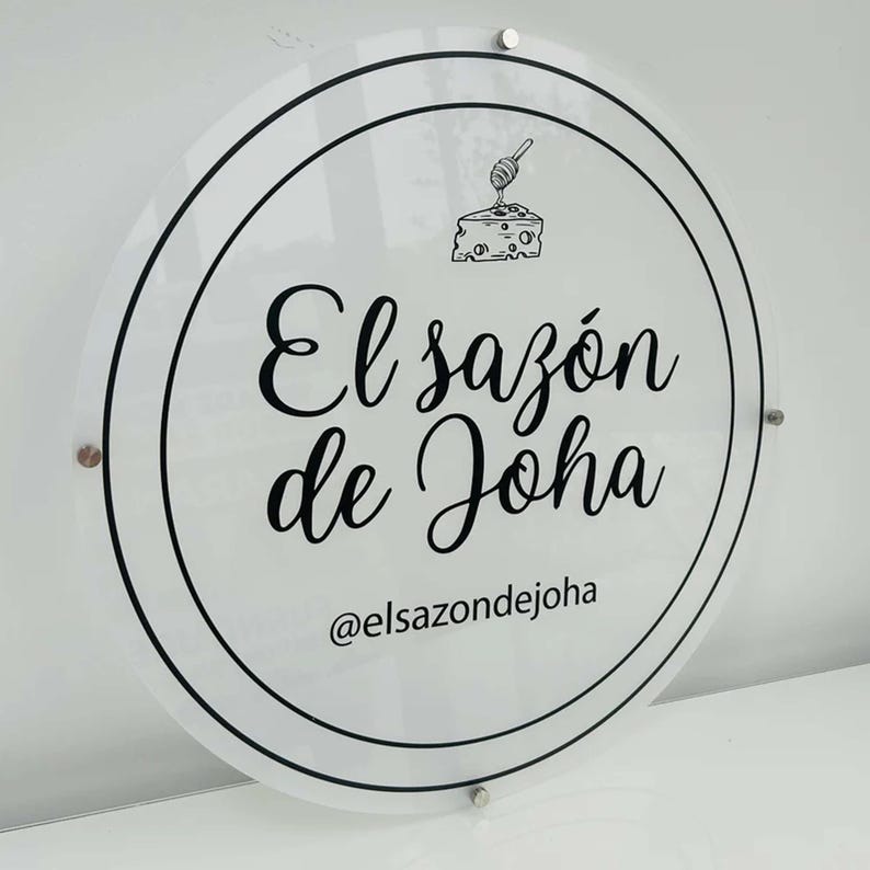 Op de afbeelding: Een wit rond bord met zwarte tekst en een afbeelding van een stuk kaas met een honinglepeltje. De tekst luidt "El saz&oacute;n de Joha" en "@elsazondejoha".
