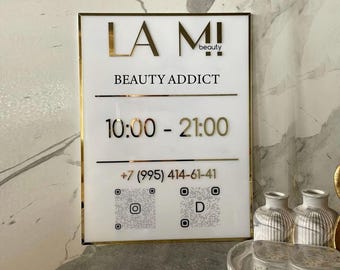 Op de afbeelding: Een rechthoekig bord met een witte achtergrond, gouden rand en tekst. Het bord toont "LA MI! beauty BEAUTY ADDICT", openingstijden "10:00 - 21:00" en een telefoonnummer. Twee QR-codes zijn ook zichtbaar.