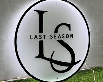 May include: A circular, backlit wall sign featuring a black border and a white translucent face. The design displays the large black letters L and S intertwined, with the text LAST SEASON printed in black capital letters across the center.