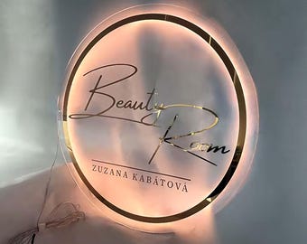 May include: A round, illuminated sign with a gold border and the text "Beauty Room" in a cursive font. The text "Zuzana Kabátová" is written below the main text.