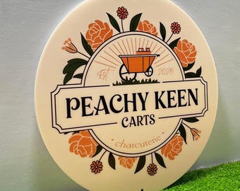 Op de afbeelding: Een ronde, crèmekleurige plaat met de tekst "PEACHY KEEN CARTS" in zwart, omringd door oranje bloemmotieven. De plaat bevat ook een kruiwagenafbeelding en het woord "charcuterie".