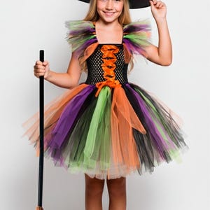 May include: A Halloween witch costume for a child. The outfit includes a black and orange striped witch hat, a black bodice with orange lacing, and a colorful tutu skirt in shades of orange, purple, green, and black. A broom completes the ensemble.