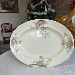 May include: Oval, off-white serving platter with a floral design featuring pink roses and blue accents. The platter is displayed on a gold-colored metal stand. The background includes a Christmas tree and a brick wall.