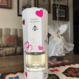 May include: A white Jasmine Blossom fragrance diffuser with a clear glass container. The packaging features pink hearts and the text "Jasmine Blossom" and "GC FRAGRANCE". The diffuser is designed to release fragrance into the air.