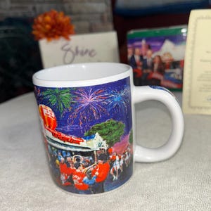 May include: White ceramic mug with a colorful illustration of a parade, fireworks, and a hot air balloon. The mug has a curved handle and a blue rim. The words "Grand Opening" are visible on the mug.