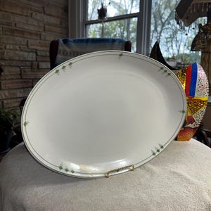 May include: Oval white serving platter with a thin green border and small green floral accents. The platter is displayed on a gold-colored metal stand. A classic piece for serving food.
