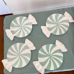 May include: Four candy-shaped plates with a mint green and white spiral design. The plates have a white, ruffled edge, resembling wrapped candies. The plates are arranged on a light blue surface.