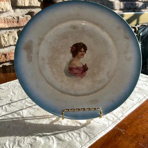 May include: A vintage porcelain plate with a blue rim and a painted image of a woman in a pink dress. The plate is on a gold stand.