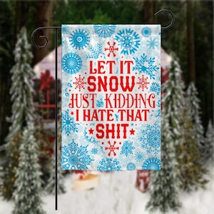 May include: A white garden flag with blue snowflakes and the text "LET IT SNOW JUST KIDDING I HATE THAT SHIT" in red. The flag is hanging on a black metal stand, with a blurred winter scene in the background.