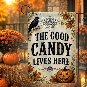 May include: A rectangular garden flag with a cream background featuring a black crow, a spiderweb, and orange autumn leaves. The text reads THE GOOD CANDY LIVES HERE in black lettering. A jack-o'-lantern is illustrated at the bottom right corner.