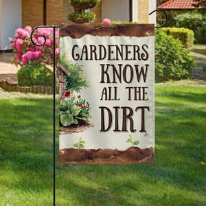 May include: A garden flag with the text "Gardeners Know All The Dirt" in brown letters. The flag has a decorative border and features a small birdhouse, plants, and flowers. The flag is displayed on a black metal stand in a grassy yard.