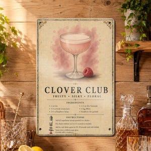 May include: A rectangular metal sign featuring a Clover Club cocktail recipe. The design includes a coupe glass filled with a pink drink, a single raspberry, and text listing ingredients like 2 oz Gin and 1/2 oz Dry Vermouth with instructions.