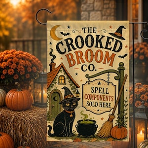 May include: A rectangular garden flag featuring a beige background with the text THE CROOKED BROOM CO. and SPELL COMPONENTS SOLD HERE. The design includes a black cat wearing a witch hat, a cauldron, a broom, a house, a crescent moon, and bats.