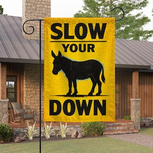 May include: Yellow garden flag with the words "SLOW YOUR DOWN" in black, with a black silhouette of a donkey. The flag is hanging outside a house.
