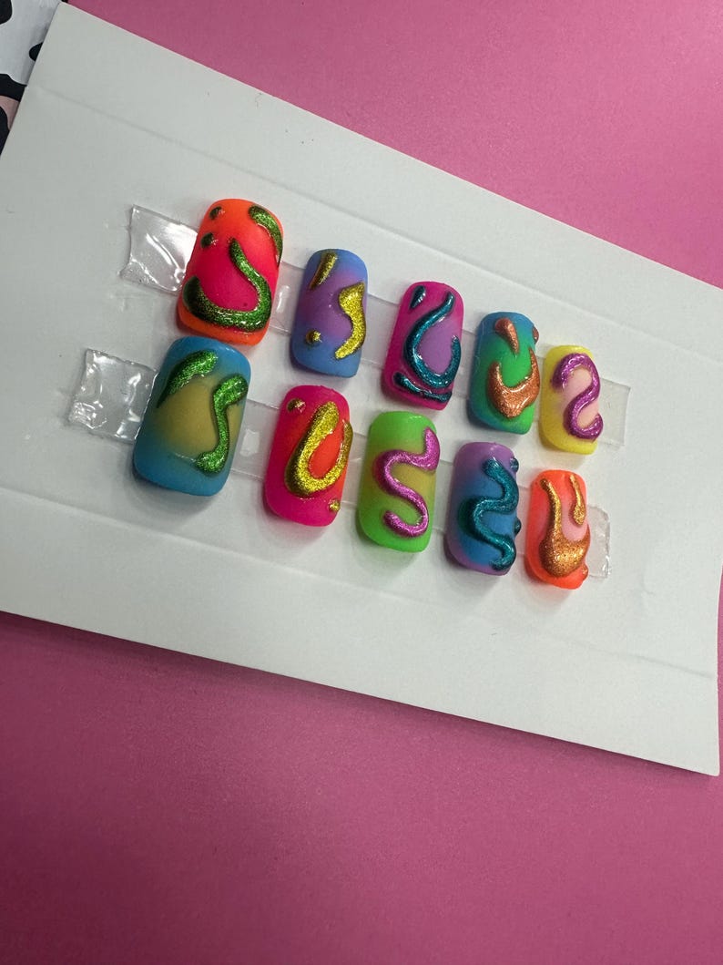May include: Set of eight colorful fake nails with abstract designs. The nails are decorated with swirls and lines in various colors, including green, yellow, orange, pink, and blue.