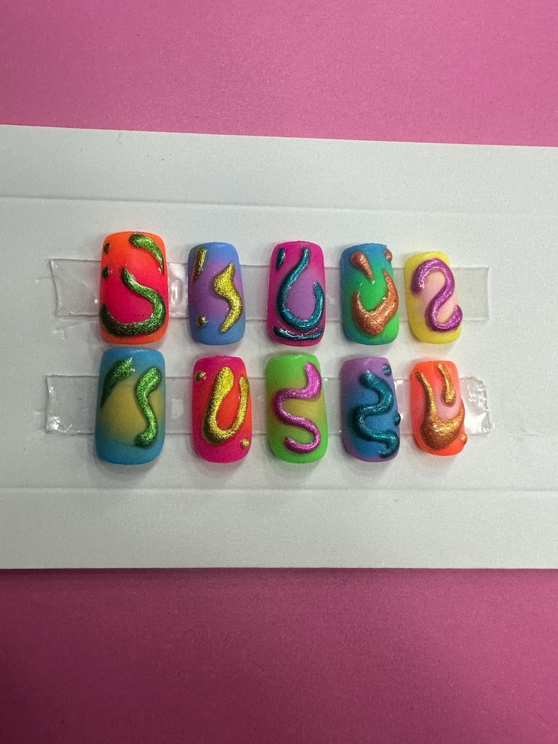 May include: Set of ten colorful fake nails with abstract designs. Each nail is a different color with a swirl design in a contrasting color. The nails are arranged in two rows of five on a white background.