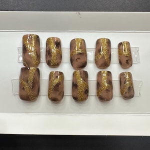 Puede incluir: Conjunto de diez uñas postizas marrones y doradas con acabado brillante. Las uñas tienen un diseño marmoleado con detalles de purpurina dorada.