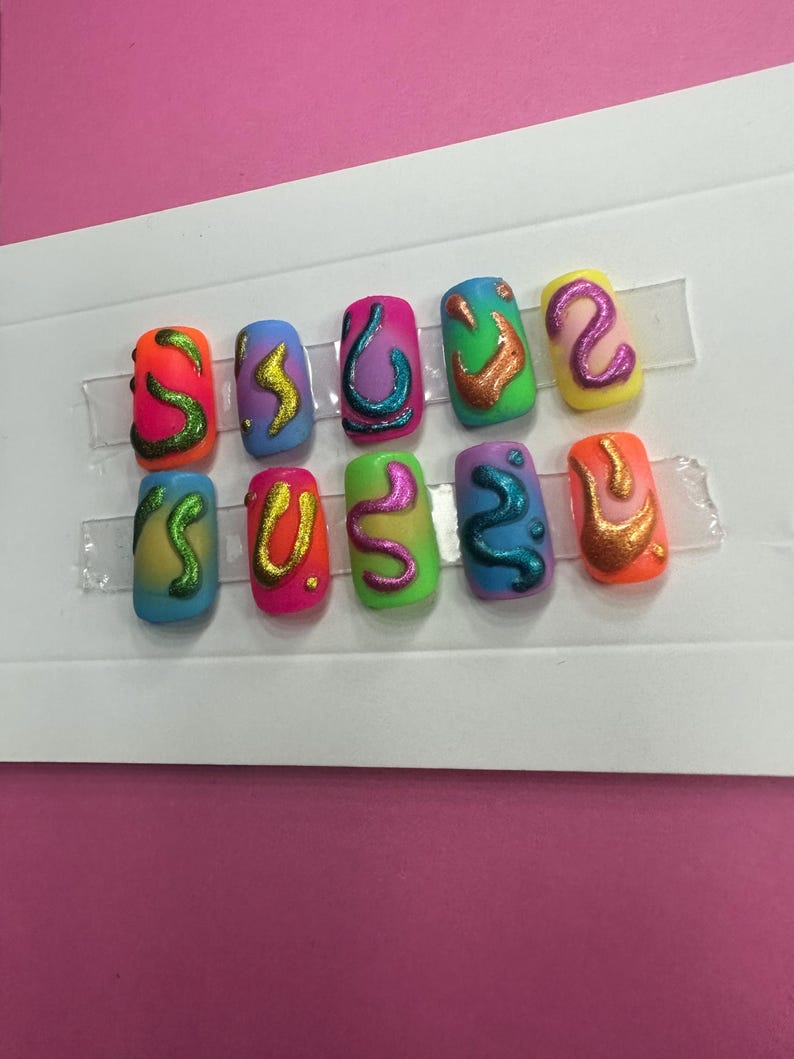 May include: Set of ten colorful fake nails with abstract designs. The nails are painted in various colors, including blue, green, yellow, orange, and pink. Each nail has a different abstract design, such as swirls, lines, and dots. The nails are arranged in two rows of five.