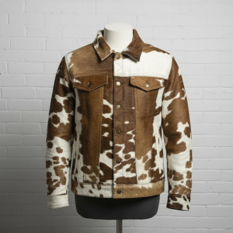 May include: A cowhide jacket with a brown and white spotted pattern. The jacket has a classic design with a collar, two chest pockets, and button closures. The jacket is displayed on a mannequin against a white brick wall.