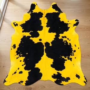 May include: A cowhide rug featuring a bright yellow base with black blotches. The rug is cut in the shape of a cowhide, with a natural, irregular outline. It is displayed on a light wooden floor.