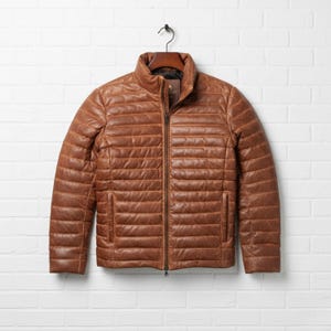 Puede incluir: Una chaqueta de cuero color canela con un patrón de costuras horizontales acolchadas, una cremallera central completa, cuello alzado y dos bolsillos laterales. La prenda se muestra en una percha, exhibiendo una textura suave y un tono uniforme.