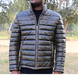 Puede incluir: Una chaqueta acolchada con cierre de cremallera y cuello alto. La chaqueta está hecha de un material similar al cuero de color marrón oscuro.