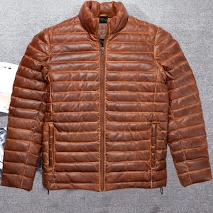 Könnte beinhalten: Eine braune, gesteppte Lederjacke mit Reißverschluss. Die Jacke hat einen Stehkragen und ein horizontales Steppmuster.