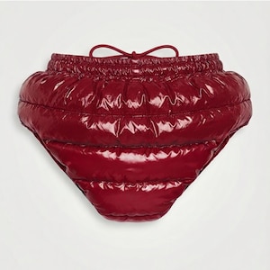Può includere: Un paio di pantaloncini trapuntati rosso scuro a vita alta con una finitura lucida simile alla vernice. Il capo presenta canali cuciti orizzontalmente, una spessa fascia elastica in vita e un cordino abbinato annodato al centro.